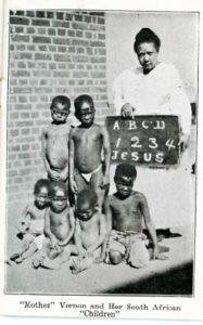 An adult woman stands next to 6 young children. The woman holds a chalkboard with the following text: A, B, C, D, 1, 2, 3, 4, Jesus.