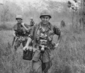 A black and white image shows a man dressed in military attire and carrying a camera case on his shoulder as well as a camera around his neck.