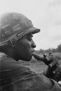 A black and white image shows a close up of a soldier who appears to be sitting and smoking a cigarette.