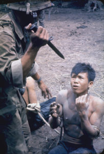 A color photograph shows a soldier standing above a man sitting on the ground. The soldier holds a knife and the seated man appears frightened.