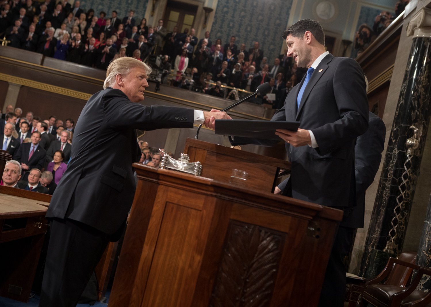 An image shows Paul Ryan standing behind a podium, reaching across the podium to shake hands with Donald Trump.