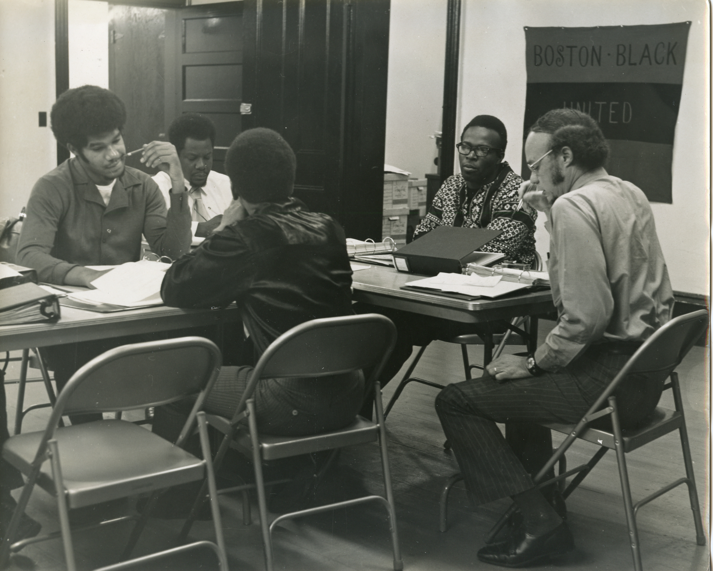 A black and white photograph shows several men sitting around a table working on papers on the table. A banner in the back reads "Boston Black United."