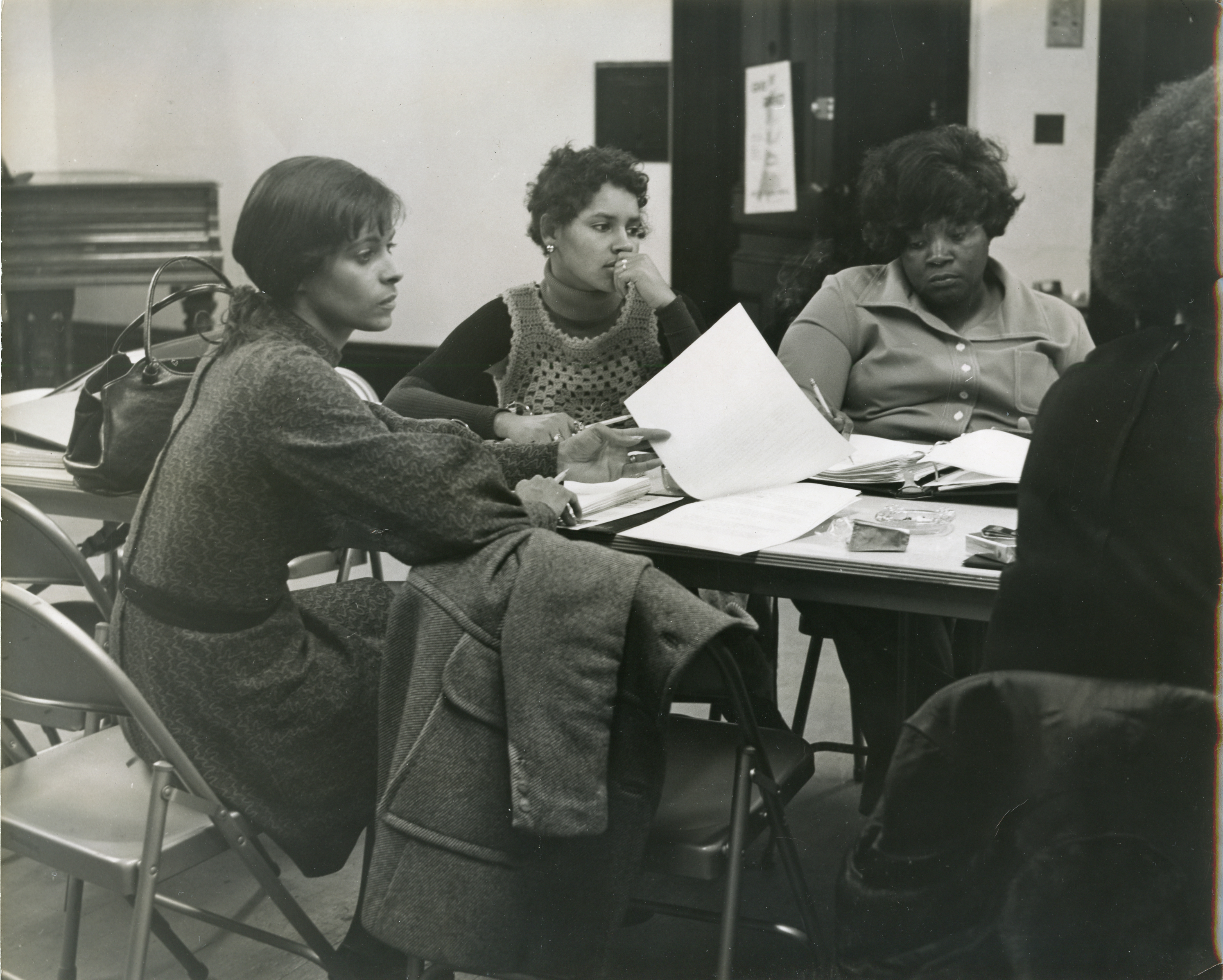 A black and white photograph shows several women sitting around a table with open binders and pieces of paper on the table.