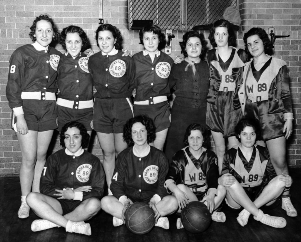 A group of women appear with one row of standing women and one row of seated women. They wear matching uniforms and two basketballs appear on the ground in front of the seated women.