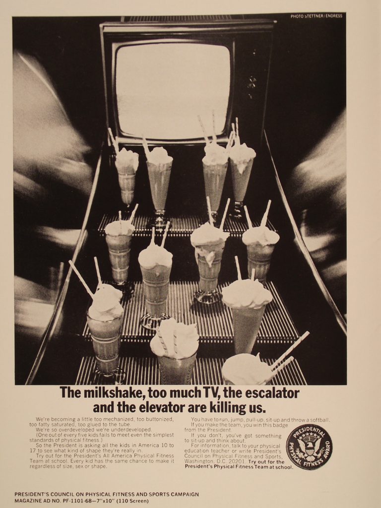 A poster shows several milkshakes and a TV set on the steps of an escalator. The poster includes the text "The milkshake, too much TV, the escalator and the elevator are killing us." The poster also has the logo of the Presidential Physical Fitness Award.
