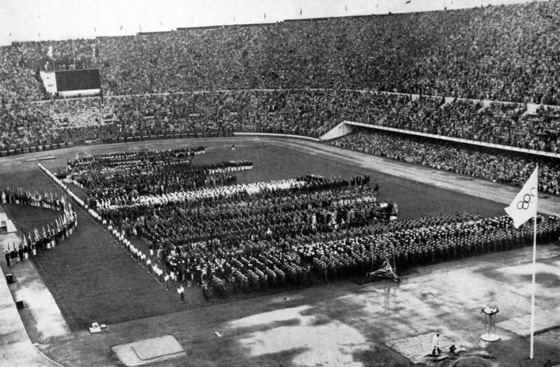 An image shows many rows of people standing in the middle of a large stadium.