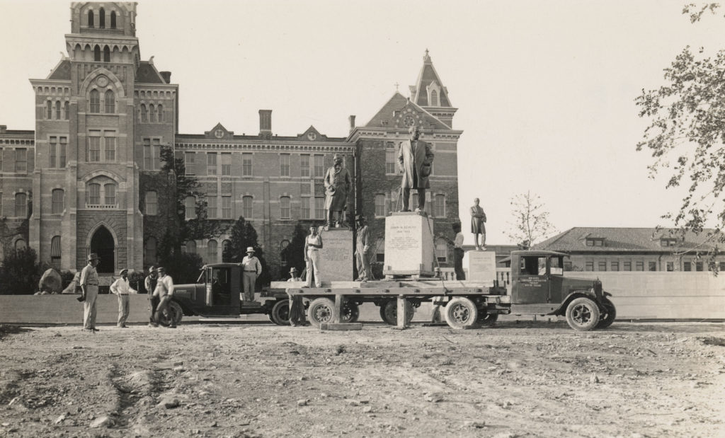 Workers stand next to a larger truck carrying three statues in front of a large university building.
