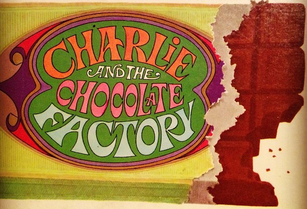 This photograph of a book cover displays a partly eaten chocolate bar has a wrapper which displays the text Charlie and the Chocolate Factory in bright colors.