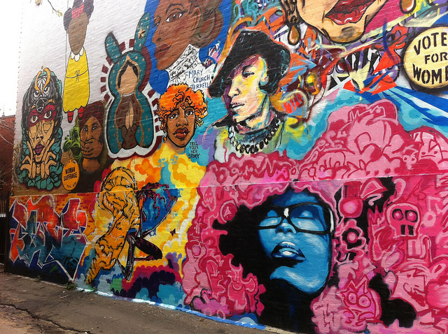 A colorful, abstract wall mural depicts women and the words "Votes for Women," "Woman Suffrage," and "Tell Your Herstory" 