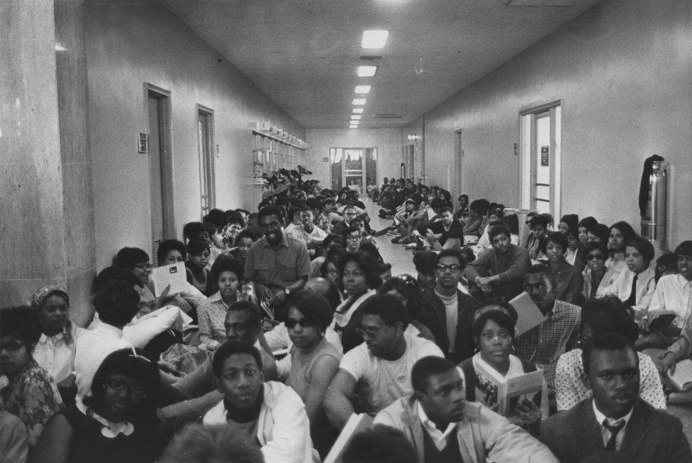 A large group of young African American men and women sit in a wide hallway, filling the space and making it difficult to pass through.