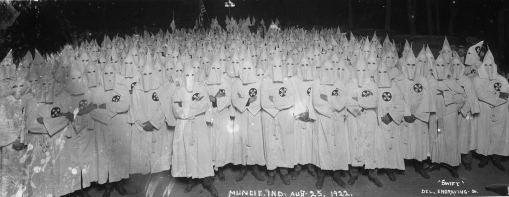 A large group of men wear white robes and hoods. The American flag is visible in the background.