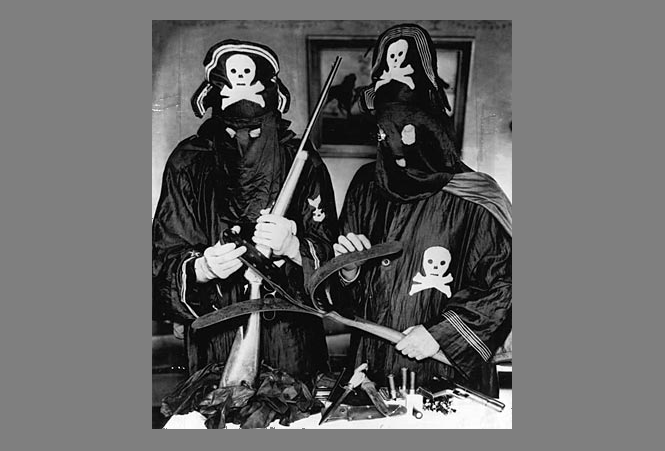 Two men wearing black robes, hoods, and hats with skulls and crossbones stand in front of a table holding weapons.