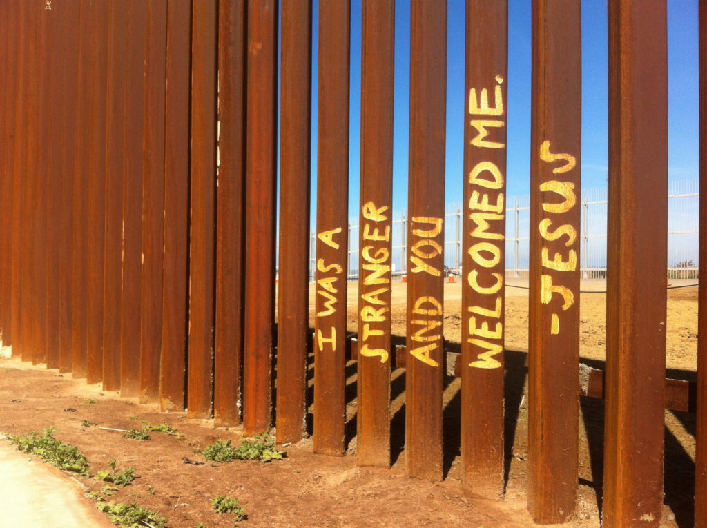 In yellow paint, the words "I was a stranger and you welcomed me - Jesus" are written on brown, rusty fence slats.