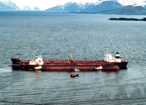 The huge ship appears in Prince William Sound. Mountains are visible in the background, and several response vehicles surround the oil tanker.