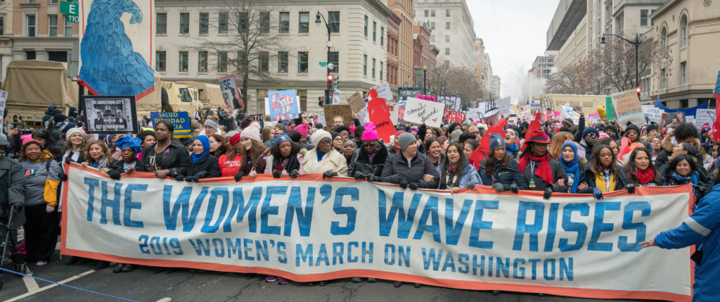 A large group of women carry many signs, including a large banner that reads "The Women's Wave Rises: 2019 Women's March on Washington."