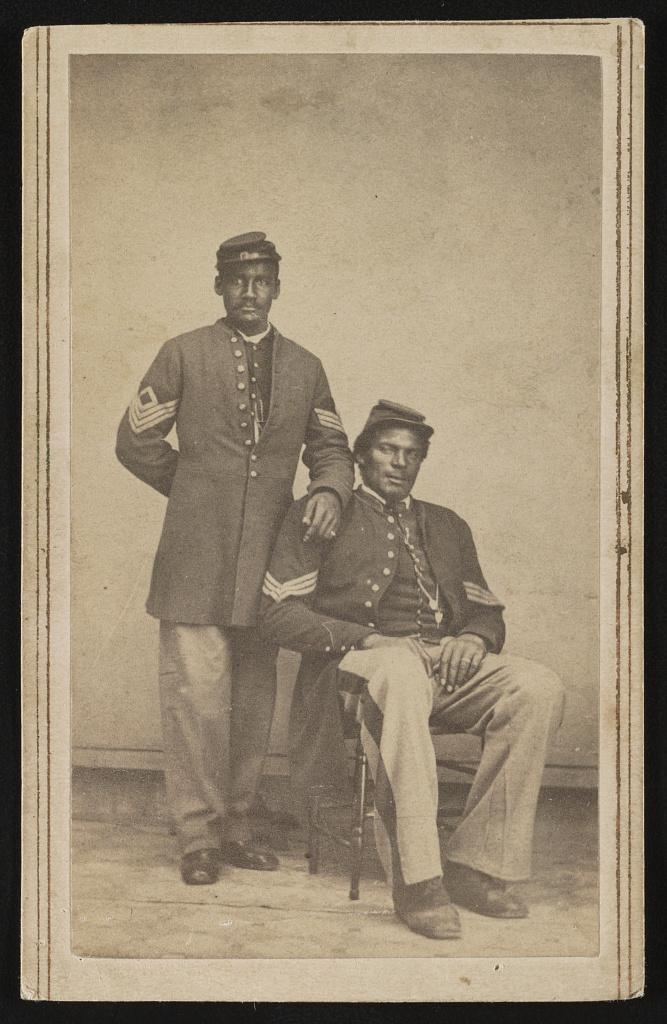 Two African American soldiers pose for a photograph in their Union Army Uniforms. One sits, and the other stands with his hand on his comrade's shoulder. 