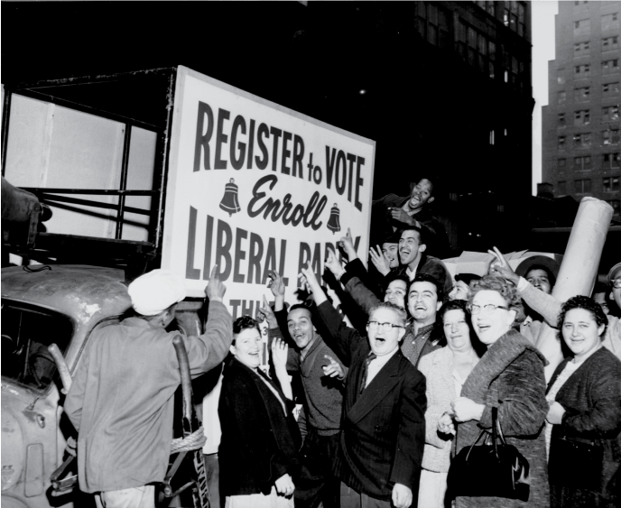 A black and white photo shows a crowd of excited men and women pointing to a sound truck with the sign "Register to Vote: Enroll Liberal Party"