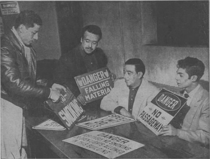Black and white photo of students at Dehon College. They sit at a table holding signs from a steel mill.