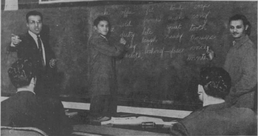 A photo of Robert Segovia, instructing a class. Two students stand at the chalkboard with Segovia.