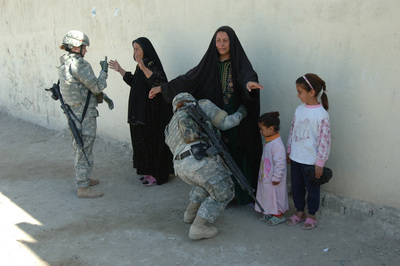 Two Iraqi women stand against a wall, arms outstretched, next to two children. Two U.S. army soldiers search the women, one patting them down.
