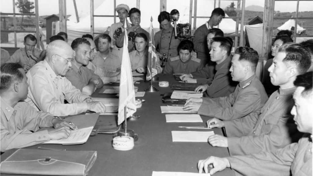 A photograph of miliary servicemen in North Korean and Unite States military uniforms sitting across from one another at a long table. with flags and stacks of papers between them.