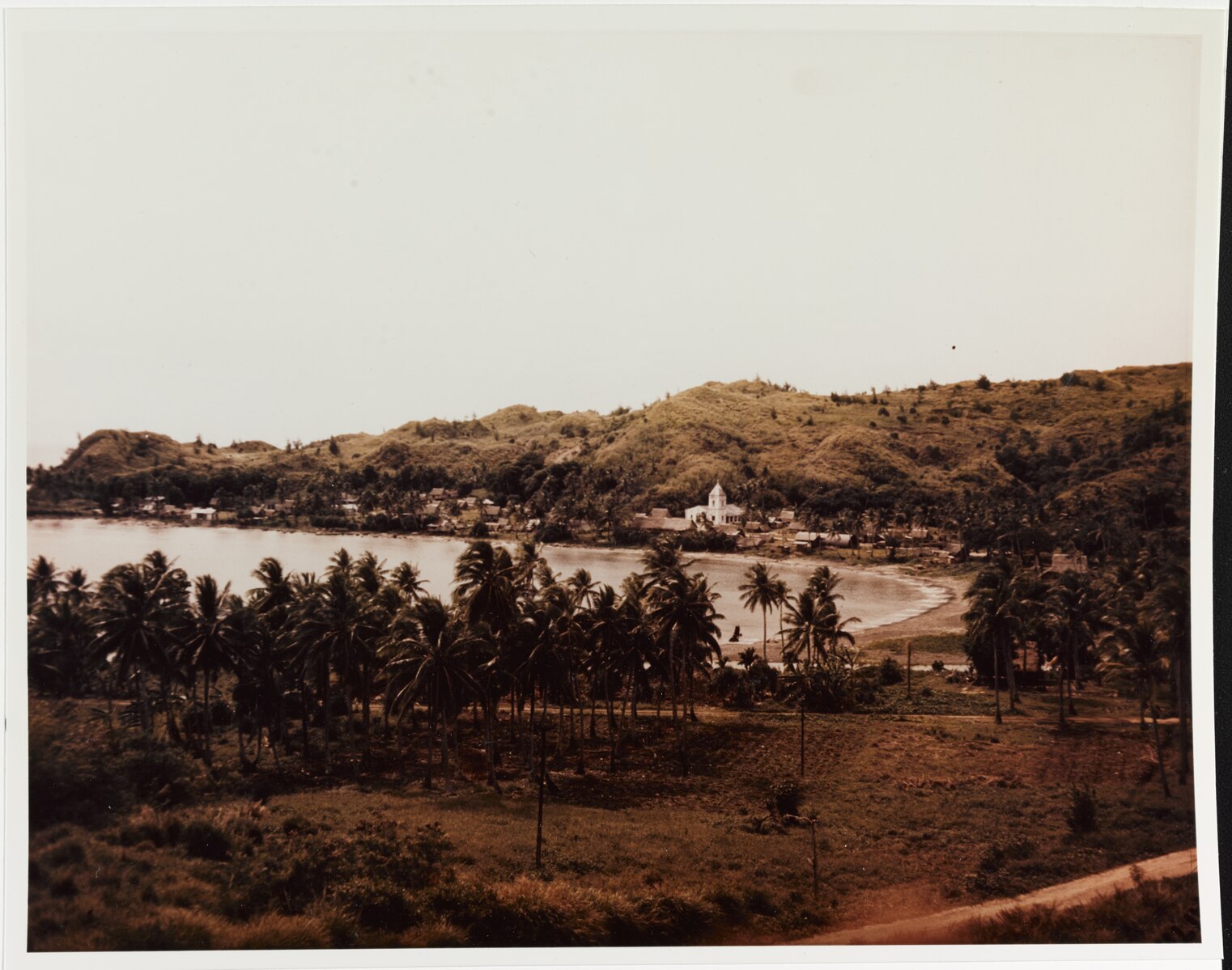 A sepia-toned photograph of Humåtak Bay and Village in Guåhan. The photo shows water surrounded by hills and palm trees and the town.