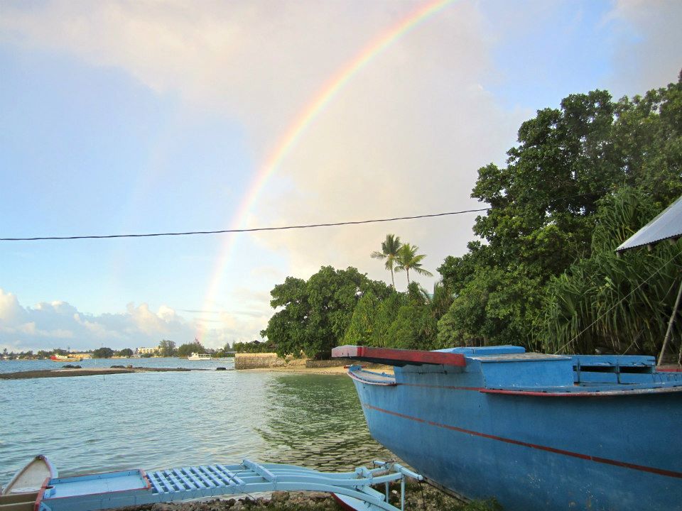 Looking out from shore at a lagoon, with a blue boat in the foreground, and a rainbow in the background.