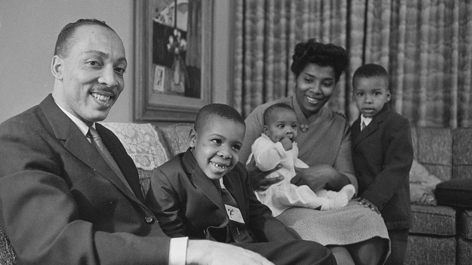 A black and white photo of Black family sitting on a couch. A man sits closest to the viewer, looking into the camera and smiling. Two small boys sit on either side of a woman who smiles at a baby in her lap. 